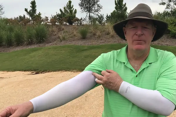 A golfer beside a bunker wearing white IceRays arm sleeves, smoothing the top band up under his shirt sleeve