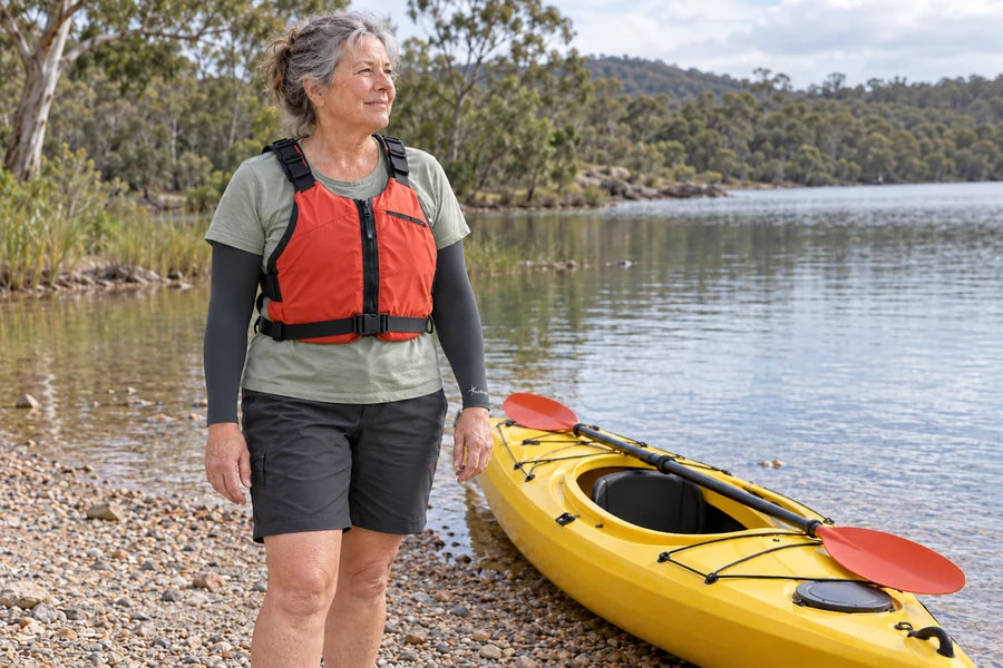 Woman wearing charcoal IceRays arm sleeves and a buoyancy vest beside a kayak at the shore.